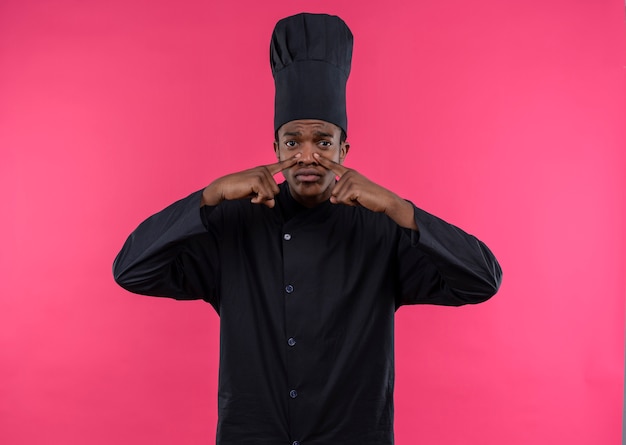 Photo gratuite jeune cuisinier afro-américain agacé en uniforme de chef ferme le nez avec les doigts des deux mains isolé sur fond rose avec copie espace