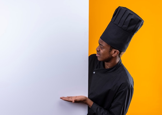 Jeune cuisinier afro-américain agacé en uniforme de chef se tient derrière un mur blanc et des points au mur isolé sur fond orange avec copie espace