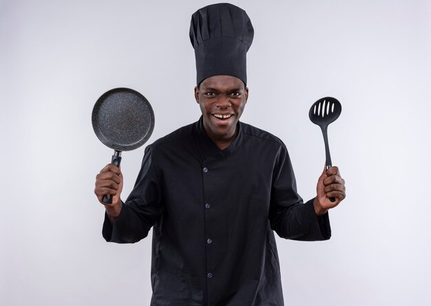 Jeune cuisinier afro-américain agacé en uniforme de chef tient une poêle et une spatule sur blanc avec copie espace