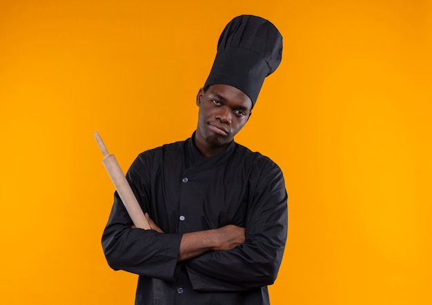 Jeune cuisinier afro-américain agacé en uniforme de chef tient le rouleau à pâtisserie avec les bras croisés sur orange avec copie espace