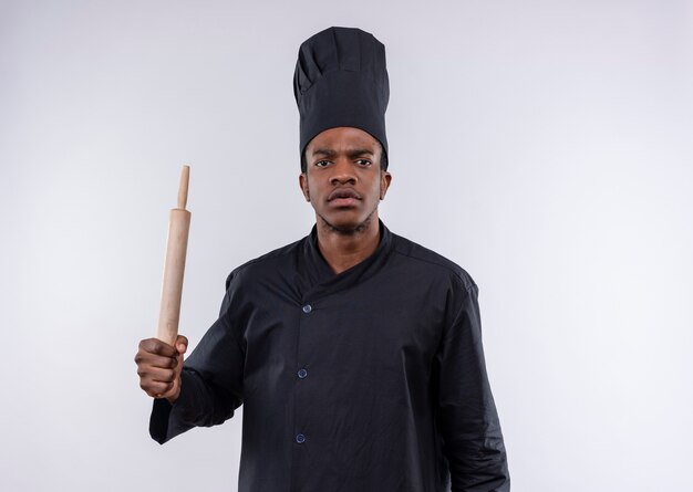 Jeune cuisinier afro-américain agacé en uniforme de chef tient le rouleau à pâtisserie isolé sur fond blanc avec copie espace