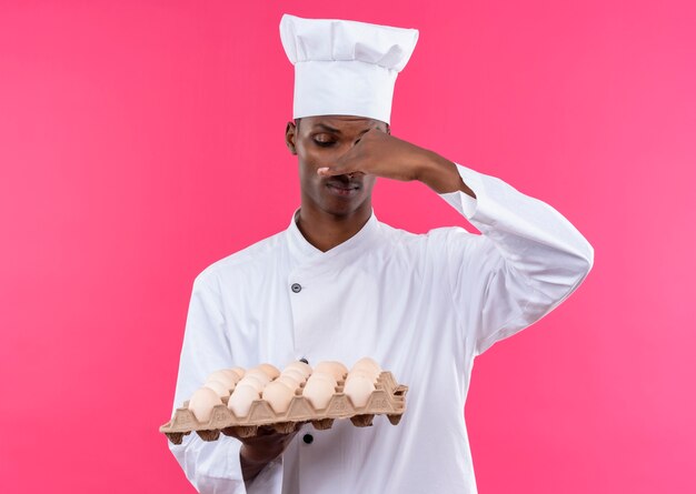 Jeune cuisinier afro-américain annpyed en uniforme de chef détient lot d'oeufs frais et ferme le nez avec la main isolé sur fond rose avec copie espace