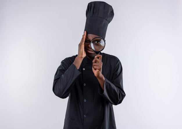 Jeune cuisinier afro-américain anxieux en uniforme de chef regarde à travers une loupe ou une loupe sur blanc avec espace de copie