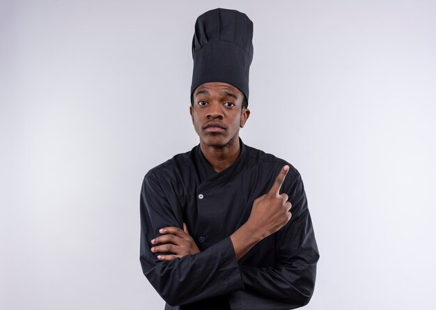 Jeune cuisinier afro-américain confiant en uniforme de chef croise les bras et pointe vers le haut isolé sur fond blanc avec copie espace