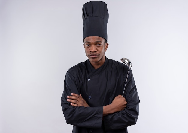 Jeune cuisinier afro-américain confiant en uniforme de chef croise les bras et tient la louche isolé sur fond blanc avec copie espace