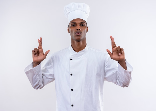 Jeune cuisinier afro-américain confiant en uniforme de chef croise les doigts des deux mains isolé sur fond blanc avec copie espace