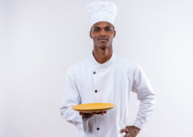 Jeune cuisinier afro-américain confiant en uniforme de chef détient une assiette vide et met la main sur la taille sur fond blanc isolé avec copie espace