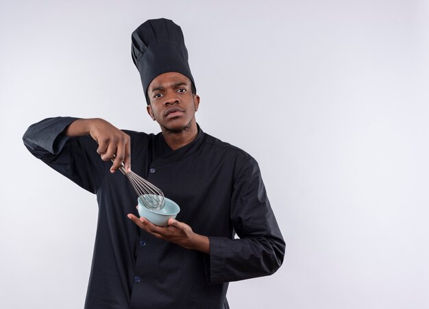 Jeune cuisinier afro-américain confiant en uniforme de chef détient bol et fouet et regarde la caméra isolée sur fond blanc avec copie espace