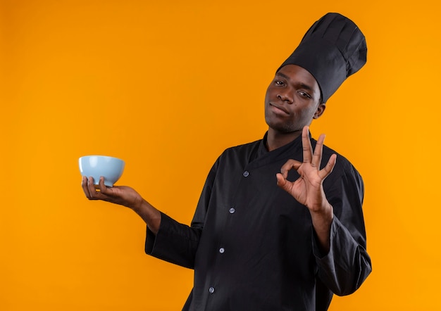 Photo gratuite jeune cuisinier afro-américain confiant en uniforme de chef détient bol et gestes ok signe de la main sur l'orange avec copie espace