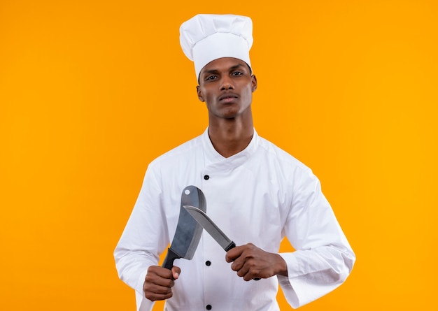 Jeune cuisinier afro-américain confiant en uniforme de chef détient des couteaux isolés sur fond orange avec copie espace