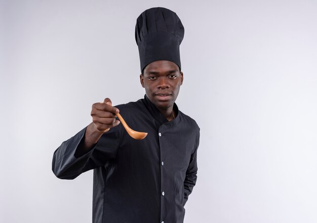 Photo gratuite jeune cuisinier afro-américain confiant en uniforme de chef détient une cuillère en bois et regarde la caméra sur blanc avec copie espace