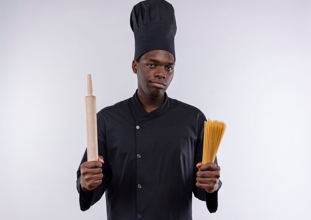 Jeune cuisinier afro-américain confiant en uniforme de chef détient un rouleau à pâtisserie et un tas de spaghettis sur blanc avec copie espace