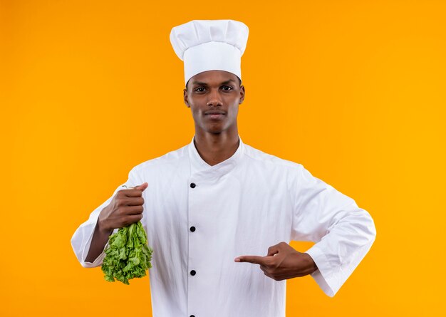 Jeune cuisinier afro-américain confiant en uniforme de chef détient la salade à l'envers et points à salade isolé sur fond orange avec copie espace