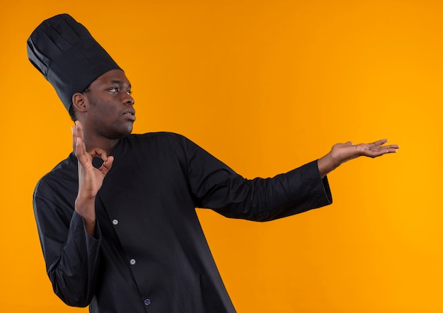 Jeune cuisinier afro-américain confiant en uniforme de chef gestes signe de la main ok et points sur le côté isolé sur l'espace orange avec copie espace