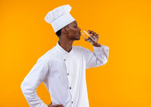 Jeune cuisinier afro-américain confiant en uniforme de chef se tient sur le côté et boit un verre d'eau isolé sur fond orange avec copie espace