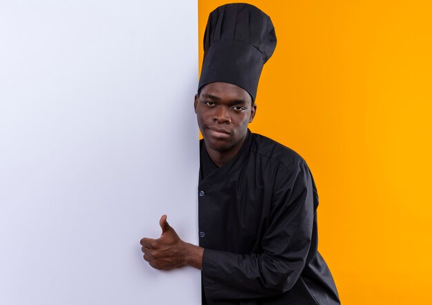 Photo gratuite jeune cuisinier afro-américain confiant en uniforme de chef se tient derrière un mur blanc et les pouces vers le haut regardant la caméra sur l'orange avec copie espace