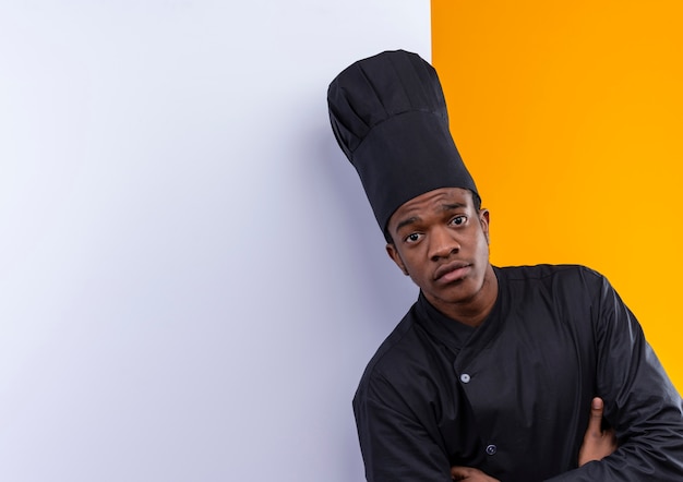 Photo gratuite jeune cuisinier afro-américain confiant en uniforme de chef se tient devant un mur blanc isolé sur fond orange avec espace de copie