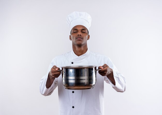 Jeune cuisinier afro-américain confiant en uniforme de chef tient une casserole avec les yeux fermés sur fond blanc isolé avec copie espace