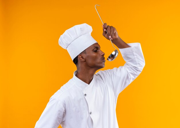 Jeune cuisinier afro-américain confiant en uniforme de chef tient la louche et fait semblant de goûter isolé sur fond orange avec copie espace