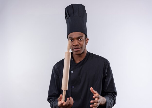 Jeune cuisinier afro-américain confiant en uniforme de chef tient le rouleau à pâtisserie tout droit sur le doigt isolé sur fond blanc avec copie espace