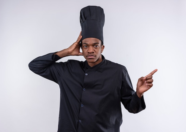 Jeune cuisinier afro-américain confiant en uniforme de chef tient la tête et pointe vers le côté isolé sur fond blanc avec copie espace