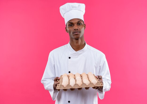 Jeune cuisinier afro-américain confiden en uniforme de chef détient lot d'oeufs frais isolé sur fond rose avec copie espace