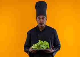 Photo gratuite jeune cuisinier afro-américain confus en uniforme de chef détient salade sur planche à découper isolé sur mur orange