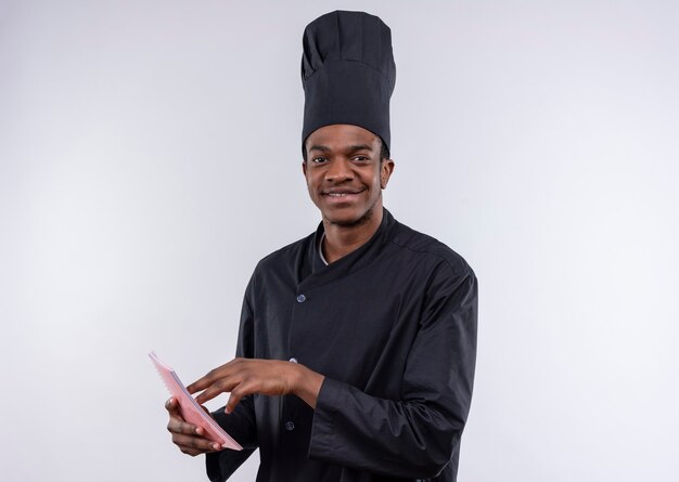 Jeune cuisinier afro-américain heureux dans les points uniformes du chef à l'ordinateur portable isolé sur mur blanc