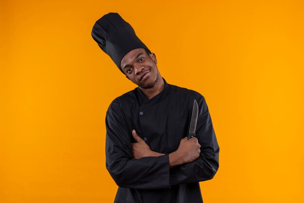 Jeune cuisinier afro-américain heureux en uniforme de chef croise les bras et tient le couteau isolé sur mur orange