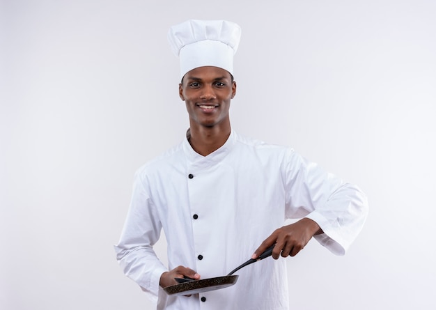 Jeune cuisinier afro-américain heureux en uniforme de chef détient une poêle et une cuillère sur un mur blanc isolé