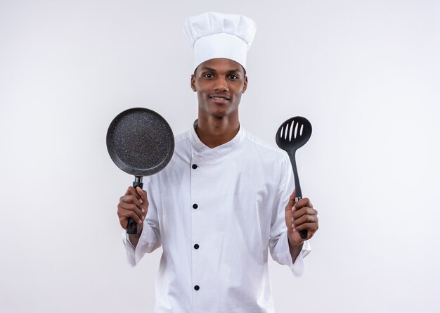 Jeune cuisinier afro-américain heureux en uniforme de chef tient une poêle et une spatule sur un mur blanc isolé