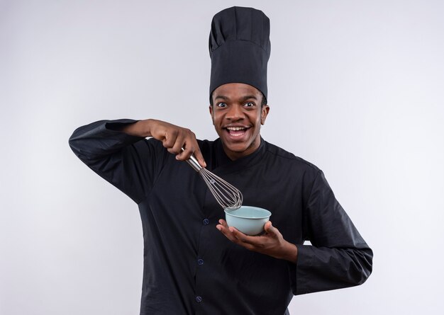 Jeune cuisinier afro-américain joyeux en uniforme de chef détient bol et fouet isolé sur mur blanc