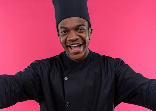 Photo gratuite jeune cuisinier afro-américain joyeux en uniforme de chef fait semblant de câliner isolé sur mur rose