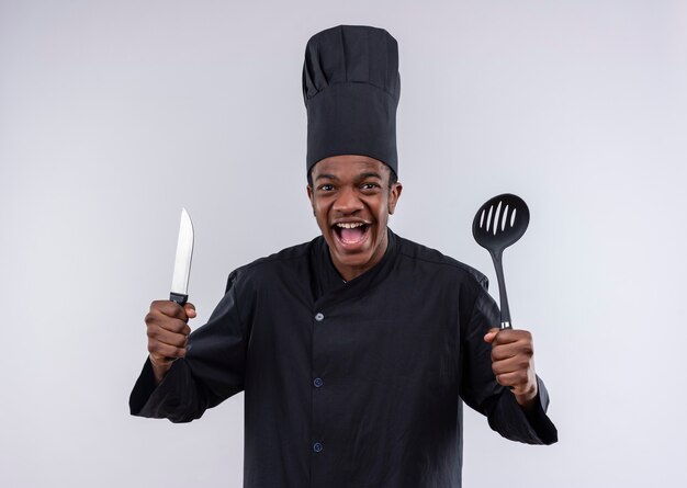 Jeune cuisinier afro-américain joyeux en uniforme de chef tient un couteau et une spatule isolé sur un mur blanc