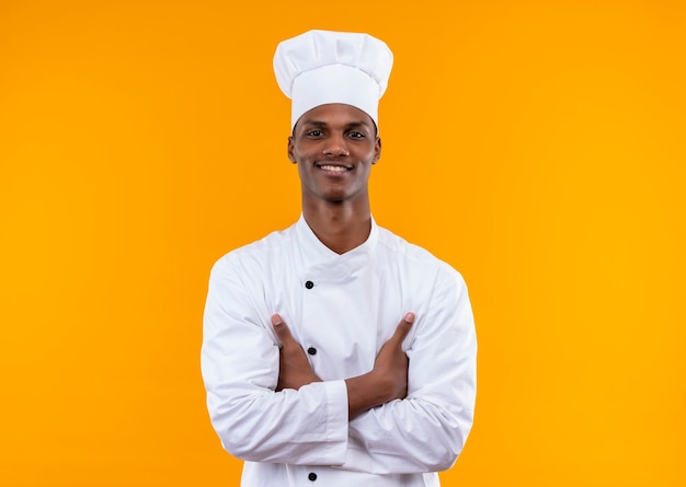 Jeune cuisinier afro-américain souriant en uniforme de chef croise les bras isolés sur mur orange