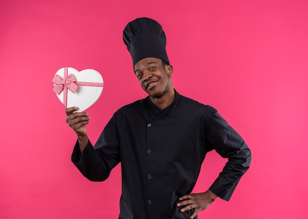 Jeune cuisinier afro-américain souriant en uniforme de chef détient une boîte en forme de coeur isolée sur un mur rose