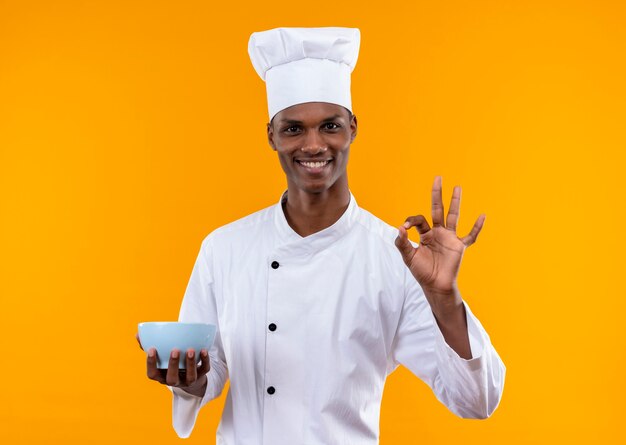 Jeune cuisinier afro-américain souriant en uniforme de chef détient bol et gestes ok avec main isolé sur mur orange