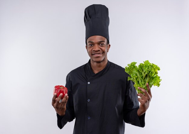 Jeune cuisinier afro-américain souriant en uniforme de chef détient poivron rouge et salade verte isolé sur mur blanc