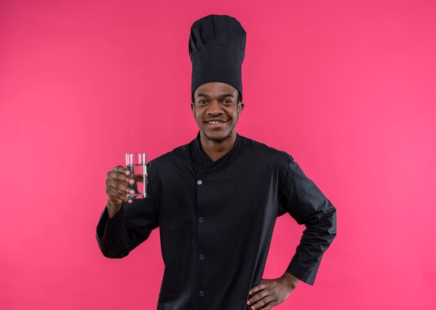 Jeune cuisinier afro-américain souriant en uniforme de chef détient un verre d'eau isolé sur un mur rose