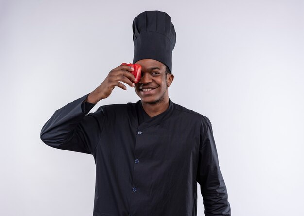 Jeune cuisinier afro-américain souriant en uniforme de chef ferme les yeux avec du poivron rouge isolé sur un mur blanc