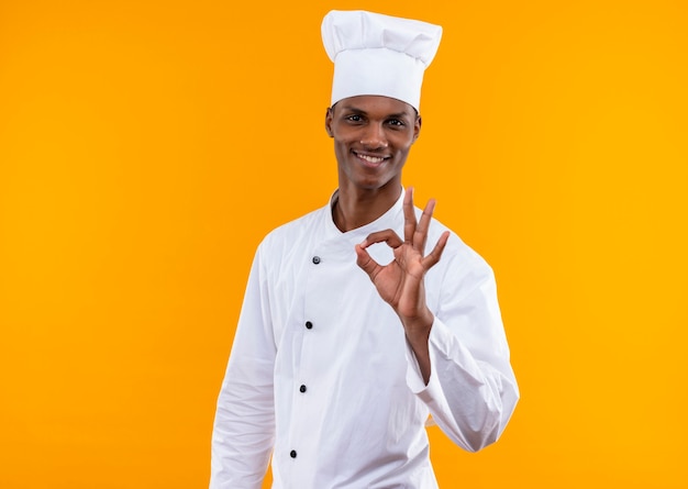 Photo gratuite jeune cuisinier afro-américain souriant en uniforme de chef gestes ok avec main isolé sur mur orange