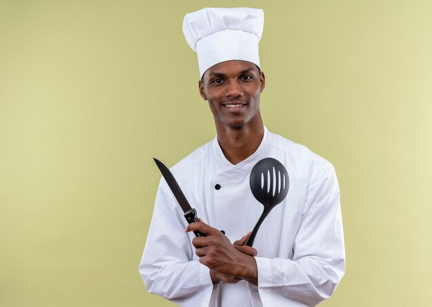 Jeune cuisinier afro-américain souriant en uniforme de chef tient un couteau et une spatule isolé sur un mur vert