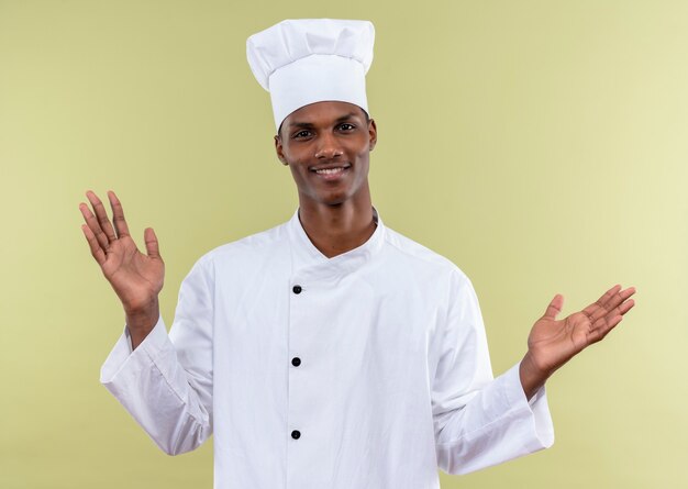 Jeune cuisinier afro-américain souriant en uniforme de chef tient les mains ouvertes isolé sur mur vert