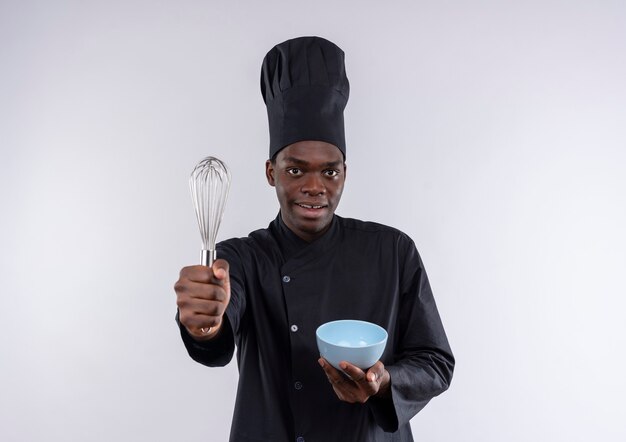 Jeune cuisinier afro-américain surpris en uniforme de chef tient un fouet et un bol sur blanc avec copie espace