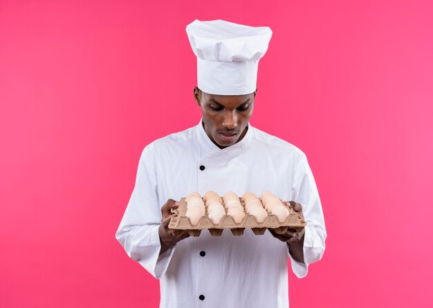 Jeune cuisinier afro-américain surpris en uniforme de chef tient et regarde lot d'oeufs frais isolé sur mur rose