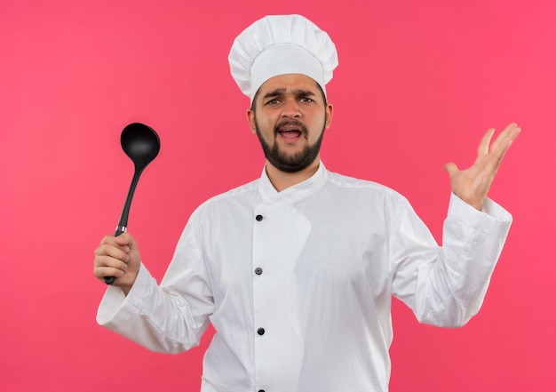 Jeune cuisinier agacé en uniforme de chef tenant une louche et montrant une main vide isolée sur un mur rose