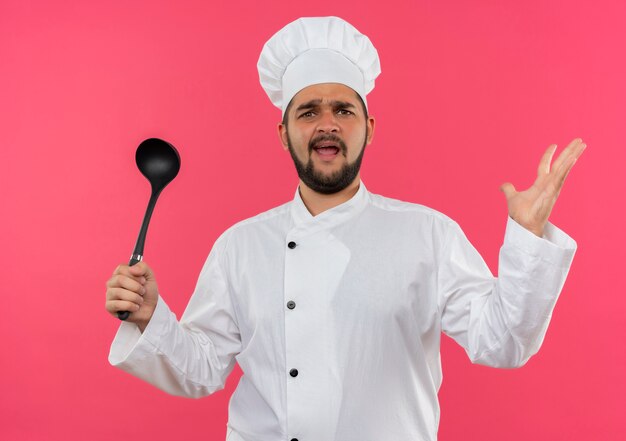 Jeune cuisinier agacé en uniforme de chef tenant une louche et montrant une main vide isolée sur un mur rose