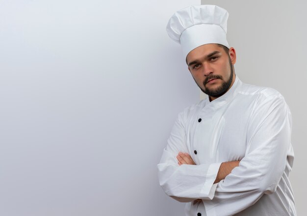 Jeune cuisinier confiant en uniforme de chef debout devant un mur blanc avec une posture fermée, isolé sur un mur blanc avec espace pour copie
