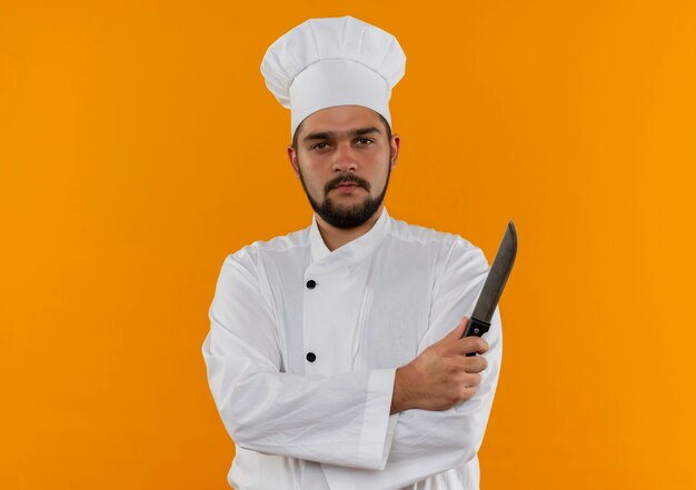 Jeune cuisinier confiant en uniforme de chef debout avec une posture fermée et tenant un couteau isolé sur un mur orange