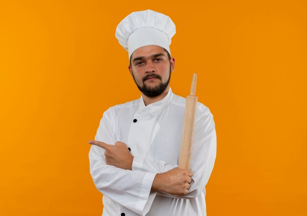 Jeune cuisinier confiant en uniforme de chef debout avec une posture fermée tenant un rouleau à pâtisserie pointant sur le côté isolé sur un mur orange avec espace de copie